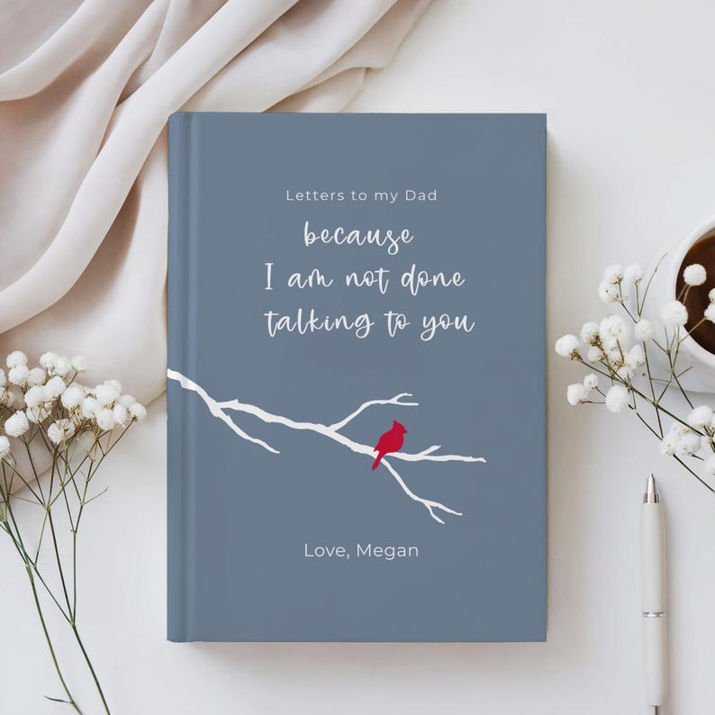 Op de afbeelding: Een hardcover journal met een blauwgrijze kaft. De tekst luidt "Letters to my Dad because I am not done talking to you." Een rode kardinaal zit op een witte takillustratie. Het journal is gepersonaliseerd met "Love, Megan."