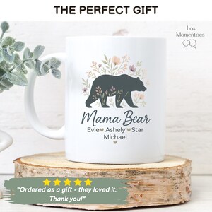 May include: White ceramic mug with a "Mama Bear" design, featuring a dark gray bear silhouette and floral accents. The mug includes names below the text. The text "THE PERFECT GIFT" is at the top.