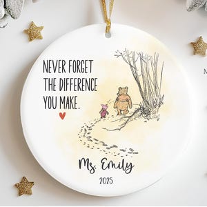 May include: A round white ceramic ornament featuring a gold ribbon hanger. The design includes a sketch of Winnie the Pooh and Piglet walking, the text "NEVER FORGET THE DIFFERENCE YOU MAKE.", a small red heart, the name "Ms. Emily", and "2025".