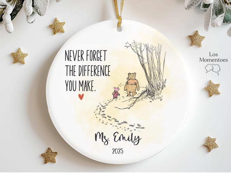 May include: A round white ceramic ornament featuring a gold ribbon hanger. The design includes a sketch of Winnie the Pooh and Piglet walking, the text "NEVER FORGET THE DIFFERENCE YOU MAKE.", a small red heart, the name "Ms. Emily", and "2025".