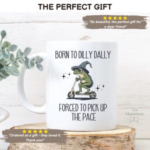 May include: White ceramic mug with a green frog wearing a witch's hat riding a scooter. The mug has the text "Born to Dilly Dally" and "Forced to Pick Up the Pace".