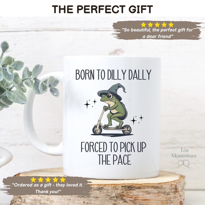 May include: White ceramic mug with a green frog wearing a witch's hat riding a scooter. The mug has the text "Born to Dilly Dally" and "Forced to Pick Up the Pace".
