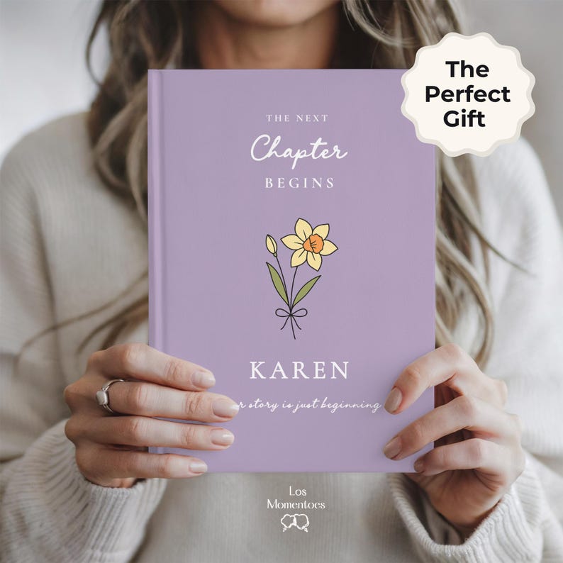 May include: A lilac journal with the text "The Next Chapter Begins" and the name "KAREN". A daffodil illustration is featured below the text. The words "The Perfect Gift" are on a white sticker in the upper right corner. The journal is held by a person.