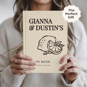 May include: A cream-colored hardcover recipe book featuring dark brown text that reads GIANNA & DUSTIN'S RECIPE BOOK from our kitchen with love. A dark brown line art illustration of a whole lemon and a sliced lemon segment is centered on the cover.