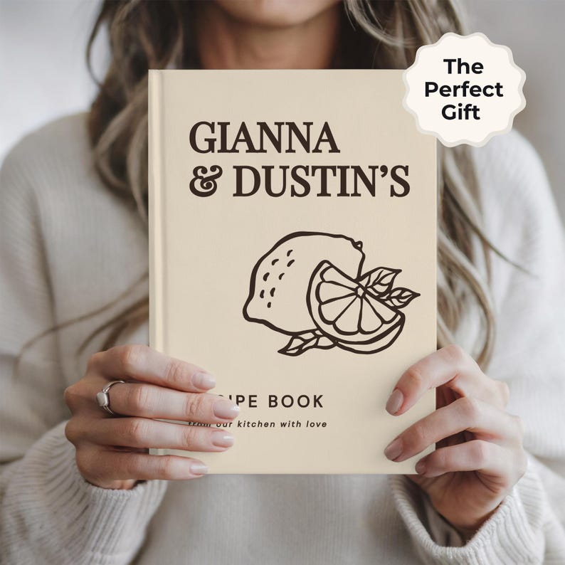 May include: A cream-colored hardcover recipe book featuring dark brown text that reads GIANNA & DUSTIN'S RECIPE BOOK from our kitchen with love. A dark brown line art illustration of a whole lemon and a sliced lemon segment is centered on the cover.