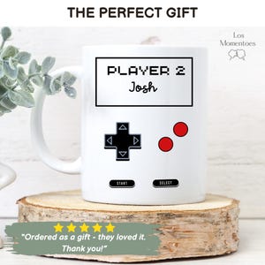 K&ouml;nnte beinhalten: Wei&szlig;e Keramik-Tasse mit einem Retro-Videospiel-Controller-Design. Die Tasse zeigt den Text "PLAYER 2 Josh" in einer pixeligen Schrift, zusammen mit einem Steuerkreuz und roten Kn&ouml;pfen. Die Tasse wird auf einer Holzscheibe pr&auml;sentiert.