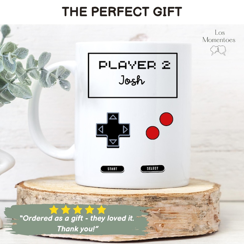 K&ouml;nnte beinhalten: Wei&szlig;e Keramik-Tasse mit einem Retro-Videospiel-Controller-Design. Die Tasse zeigt den Text "PLAYER 2 Josh" in einer pixeligen Schrift, zusammen mit einem Steuerkreuz und roten Kn&ouml;pfen. Die Tasse wird auf einer Holzscheibe pr&auml;sentiert.