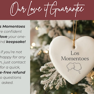 May include: A white heart-shaped ceramic ornament hanging by a gold string. The ornament features the text Los Momentoes above two cloud-shaped thought bubbles. The background shows a blurred Christmas tree with soft, warm, out-of-focus lights.