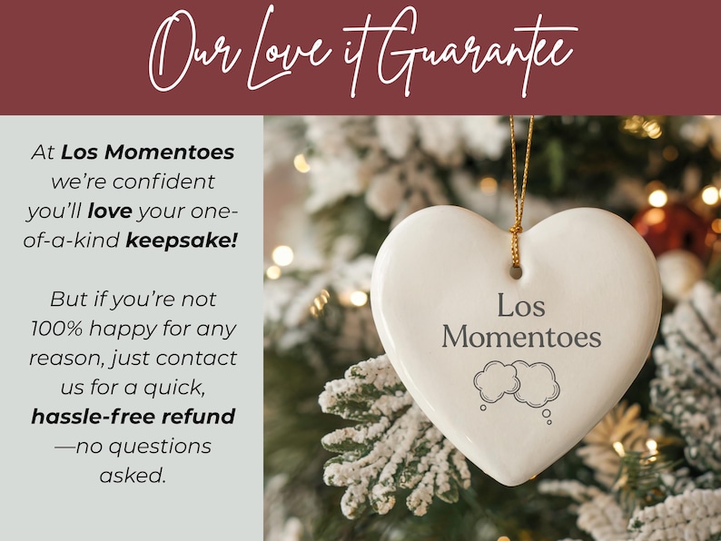 May include: A white heart-shaped ceramic ornament hanging by a gold string. The ornament features the text Los Momentoes above two cloud-shaped thought bubbles. The background shows a blurred Christmas tree with soft, warm, out-of-focus lights.