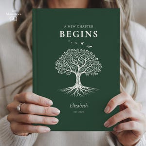 May include: A dark green journal with the words "A New Chapter Begins" and a white tree illustration. The name "Elizabeth" and the year "EST 2026" are printed below the tree. The journal is held by a person.