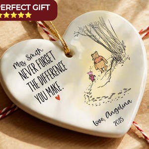 May include: A heart-shaped white ceramic ornament featuring a gold hanging string. The text reads: Mrs. Smith NEVER FORGET THE DIFFERENCE YOU MAKE. Love, Angelina 2025. An illustration shows Winnie the Pooh and Piglet walking near a tree.