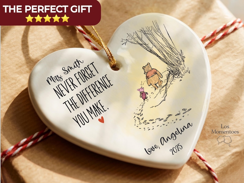 May include: A heart-shaped white ceramic ornament featuring a gold hanging string. The text reads: Mrs. Smith NEVER FORGET THE DIFFERENCE YOU MAKE. Love, Angelina 2025. An illustration shows Winnie the Pooh and Piglet walking near a tree.