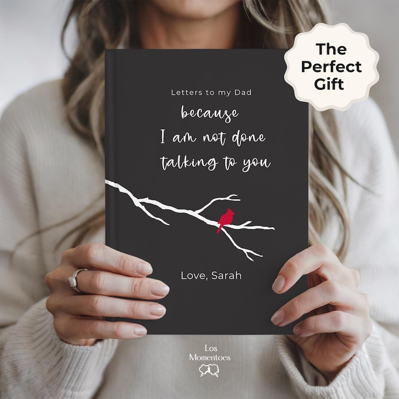 Op de afbeelding: Een zwart notitieboek met de tekst "Letters to my Dad" en de quote "because I am not done talking to you." Een witte tak met een rode vogel is afgebeeld op de omslag. Het notitieboek is gelabeld "The Perfect Gift."