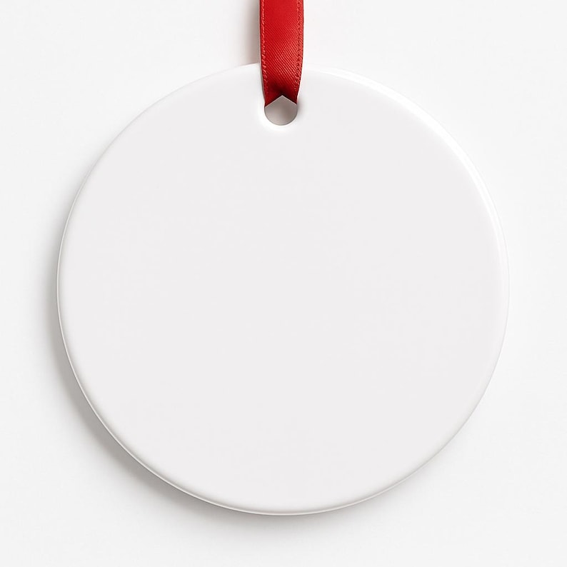 May include: A blank, white ceramic ornament with a round shape, perfect for customization. A red ribbon is threaded through a hole at the top for hanging. The ornament is smooth and glossy, ideal for holiday decorations or personalized gifts.