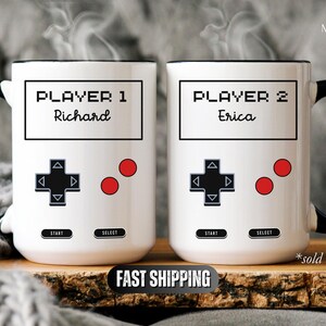 K&ouml;nnte beinhalten: Zwei wei&szlig;e Keramikbecher mit schwarzen Griffen, mit einem Videospiel-Controller-Design. Auf einem Becher steht "Player 1 Richard", auf dem anderen "Player 2 Erica". Rote Kn&ouml;pfe und ein schwarzes Steuerkreuz sind ebenfalls sichtbar.