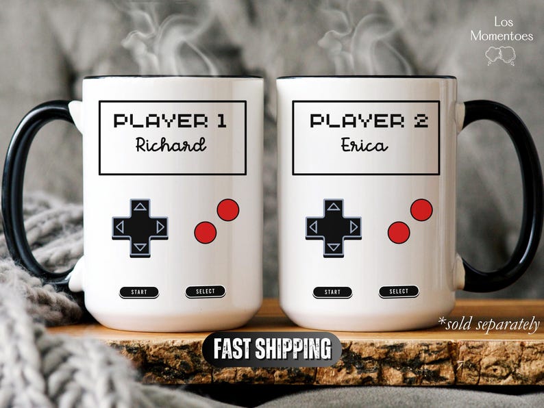 K&ouml;nnte beinhalten: Zwei wei&szlig;e Keramikbecher mit schwarzen Griffen, mit einem Videospiel-Controller-Design. Auf einem Becher steht "Player 1 Richard", auf dem anderen "Player 2 Erica". Rote Kn&ouml;pfe und ein schwarzes Steuerkreuz sind ebenfalls sichtbar.
