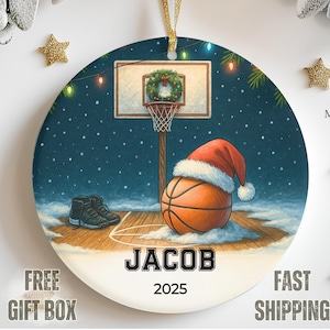 May include: Round Christmas ornament featuring a basketball with a Santa hat, a basketball hoop, and a pair of sneakers. The ornament has the name "JACOB" and the year "2025" printed on it. The background is a snowy basketball court.