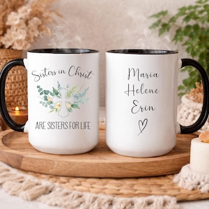 May include: Two white ceramic mugs with black handles and rims. One mug reads "Sisters in Christ ARE SISTERS FOR LIFE" with a cross and floral design. The other mug has names "Maria Helene Erin" and a heart.