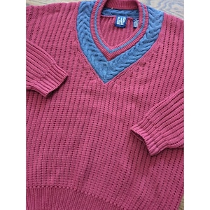 May include: A maroon V-neck sweater with a blue cable knit design around the neckline. The sweater has long sleeves with ribbed cuffs and a ribbed hem. The Gap logo is visible on the label.