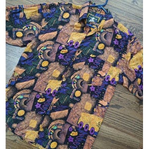 May include: A short-sleeved button-down shirt with a vibrant, abstract pattern. The design features geometric shapes, swirls, and floral elements in shades of brown, yellow, purple, and blue on a black background. The shirt is hanging on a blue hanger.