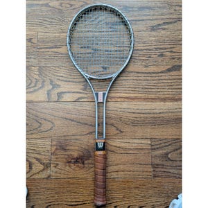 May include: A vintage Wilson tennis racket with a silver frame and a tightly strung net. The handle is wrapped in brown leather, and the grip features the Wilson logo. The racket has a classic design, perfect for tennis enthusiasts.