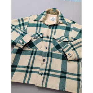 May include: A cream and dark green plaid button-up jacket featuring a pointed collar, two chest pockets with button flaps, and long sleeves with buttoned cuffs. The interior is lined with a beige sherpa material and includes a Woolrich label.