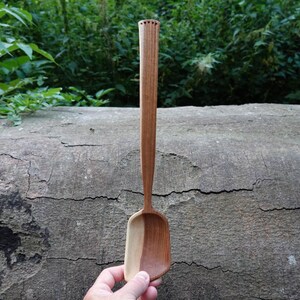 May include: A handcrafted wooden spoon with a long handle and a rectangular scoop. The spoon is made of two tones of wood, light and dark brown. The handle has a decorative carved top. The spoon is held against a textured, gray log.