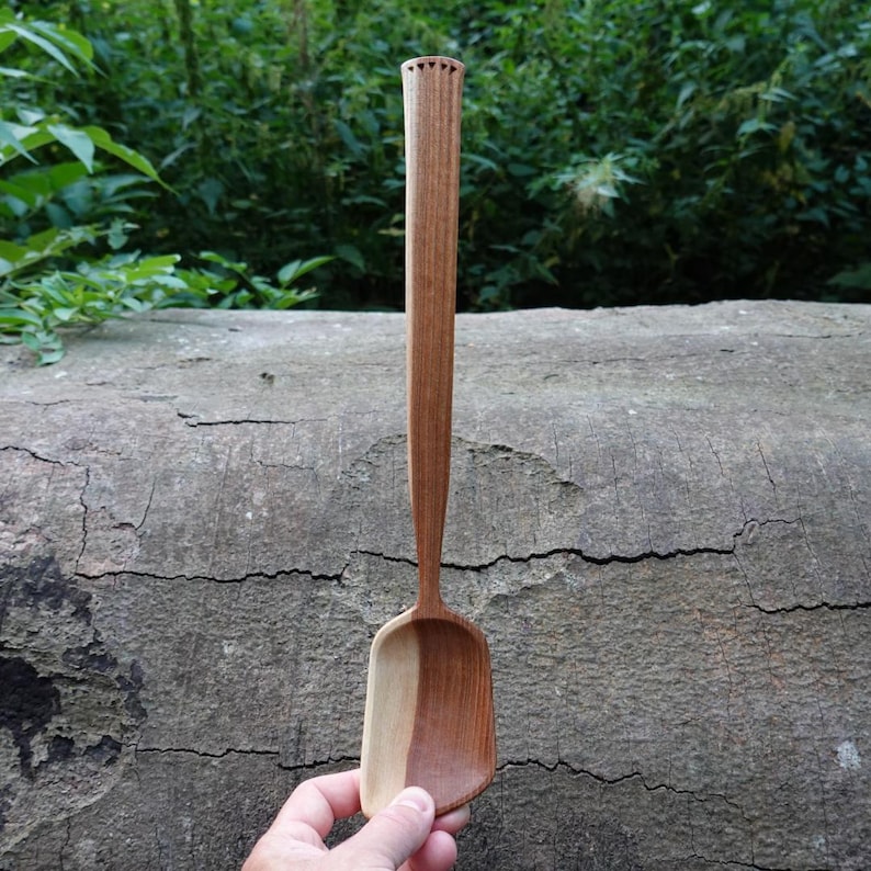 May include: A handcrafted wooden spoon with a long handle and a rectangular scoop. The spoon is made of two tones of wood, light and dark brown. The handle has a decorative carved top. The spoon is held against a textured, gray log.