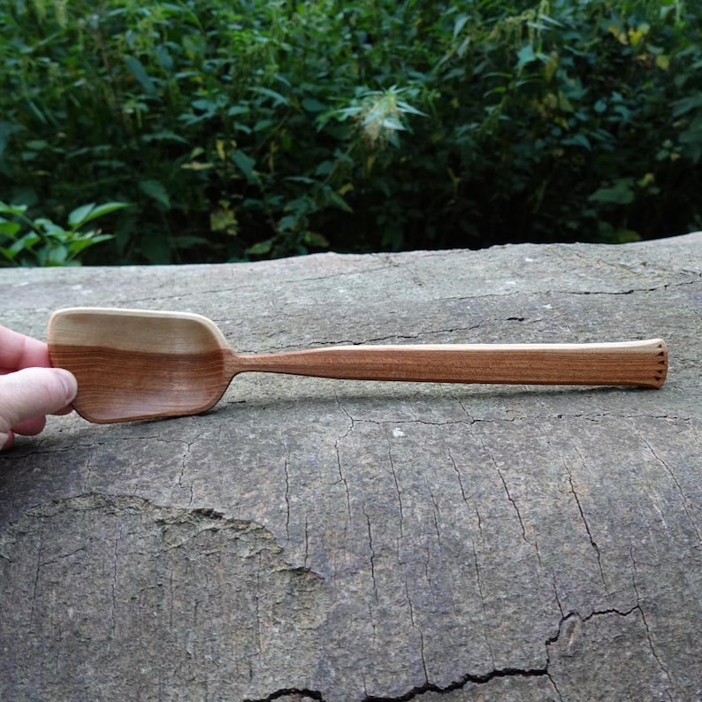 May include: A handcrafted wooden spoon with a rectangular bowl and a long, slender handle. The spoon is made of two tones of wood, with a lighter color on the bowl and a darker color on the handle. The handle has a decorative carved design at the end.