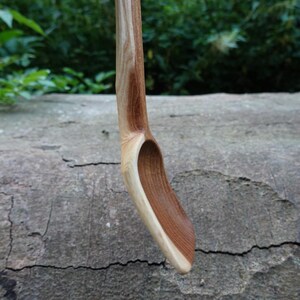 May include: A handcrafted wooden spoon with a curved handle and a deep, oval bowl. The spoon is made of light and dark brown wood, showcasing the natural grain. The spoon is designed for cooking and serving.