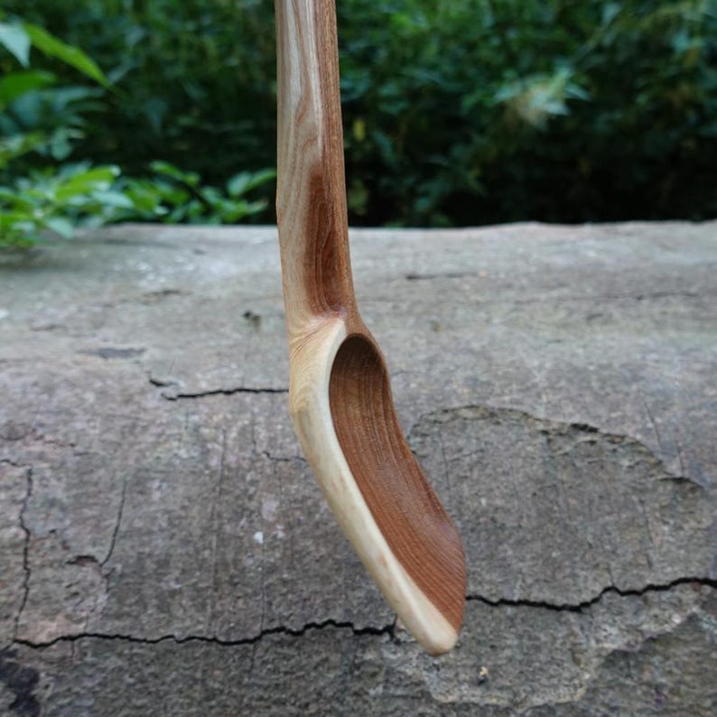 May include: A handcrafted wooden spoon with a curved handle and a deep, oval bowl. The spoon is made of light and dark brown wood, showcasing the natural grain. The spoon is designed for cooking and serving.