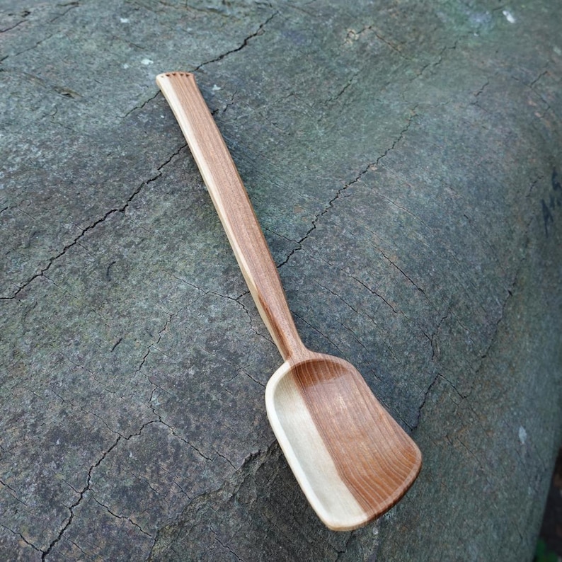 May include: A handcrafted wooden spoon with a rectangular bowl and a long, slender handle. The spoon is made of light and dark brown wood, with a natural grain pattern. The handle has a series of small notches at the end.