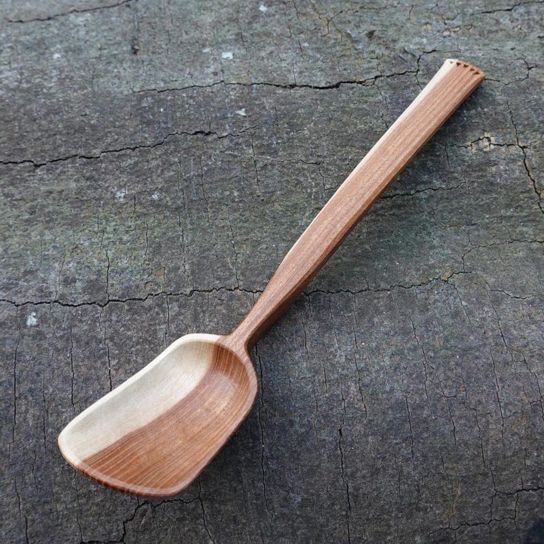 May include: A handcrafted wooden spoon with a long handle and a wide, curved bowl. The spoon is made of light and dark brown wood, with a smooth, polished finish. The handle has a decorative carved detail at the end. A kitchen utensil.