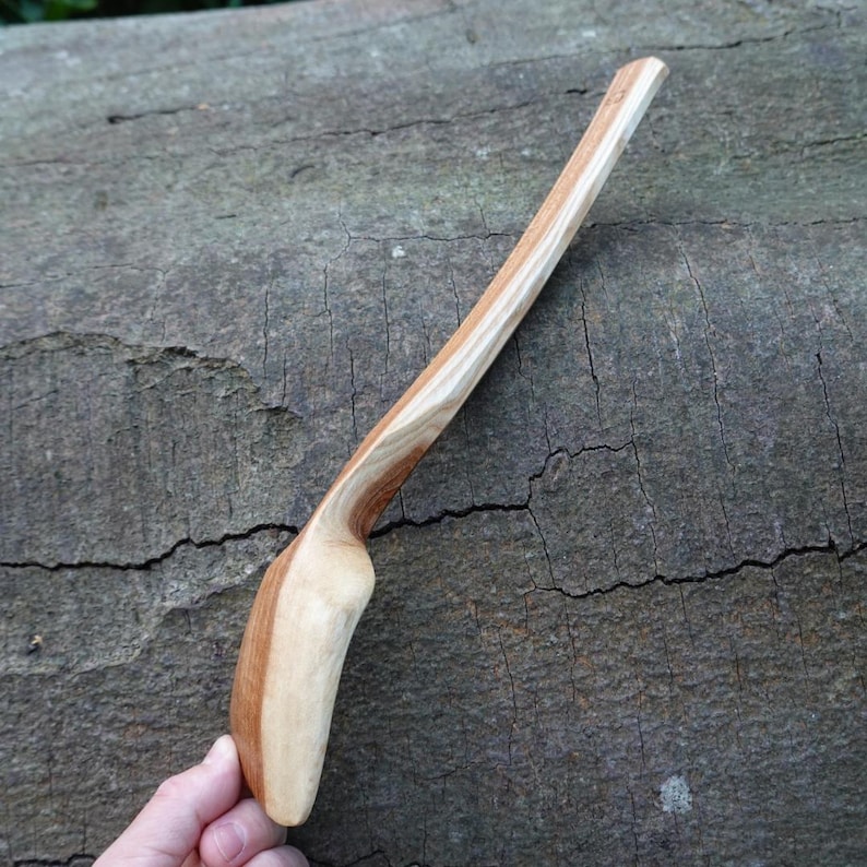 May include: A handcrafted wooden spoon with a light and dark wood grain pattern. The spoon has a curved handle and a rounded bowl. The spoon is held by a hand, showcasing the natural wood tones and craftsmanship. The spoon is approximately 12 inches long.