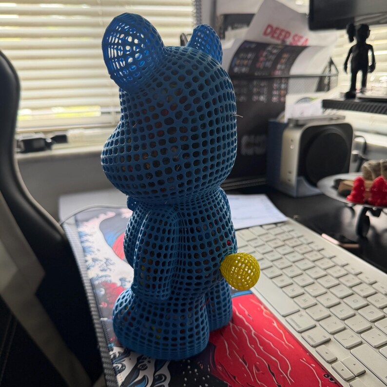 May include: A blue, open-weave bear figurine with a yellow ball attached. The bear has a rounded shape with visible holes throughout its body. The figurine is on a desk with a keyboard and other items in the background.