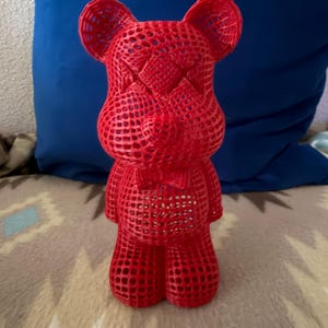 May include: A vibrant red, open-weave bear figurine. The bear has a bow tie and a textured surface. The figurine is standing upright. The bear is a decorative item.