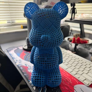 May include: A blue, open-weave bear figurine with a bow tie. The bear has a lattice-like design, with visible interior details. The figurine is standing on a desk with a keyboard and other items in the background.