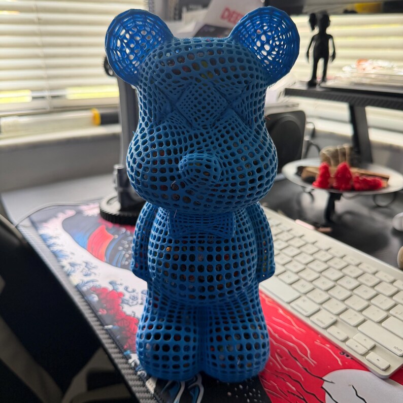 May include: A blue, open-weave bear figurine with a bow tie. The bear has a lattice-like design, with visible interior details. The figurine is standing on a desk with a keyboard and other items in the background.