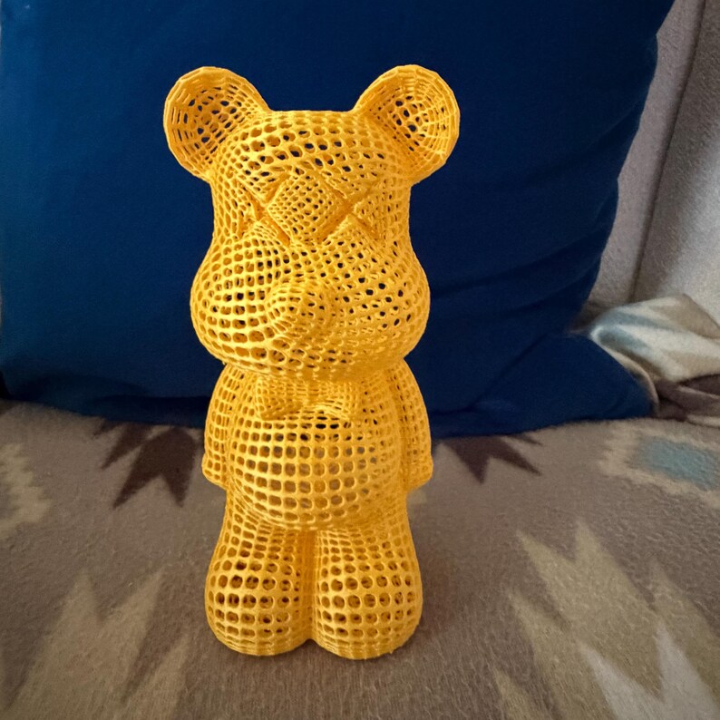 May include: A yellow, open-weave bear figurine with a distinctive design. The bear has a bow tie and a cross-shaped pattern on its face. The figurine is made of a mesh-like material, giving it a unique, modern look. The bear is standing upright.