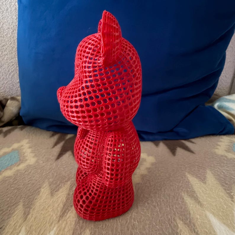May include: A vibrant red, open-weave sculpture of a bear. The bear is standing upright, with a rounded head, ears, and body. The sculpture has a lattice-like structure, creating a see-through effect. The bear is set against a blue pillow and patterned blanket.