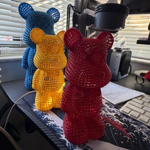 May include: Three colorful bear-shaped decorative objects. The bears are red, yellow, and blue, with a lattice design. They are standing on a desk with a keyboard and other office supplies.