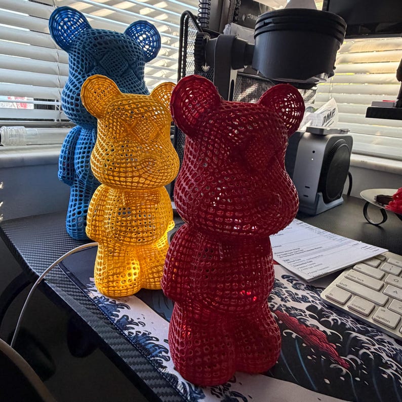 May include: Three colorful bear-shaped decorative objects. The bears are red, yellow, and blue, with a lattice design. They are standing on a desk with a keyboard and other office supplies.