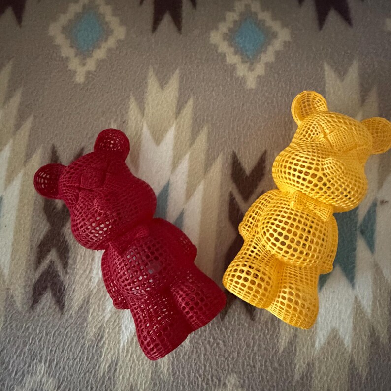May include: Two decorative bear figurines. One is red, the other is yellow. Both are made with a lattice design, giving them a unique, open appearance. They are placed on a patterned, gray and white blanket.