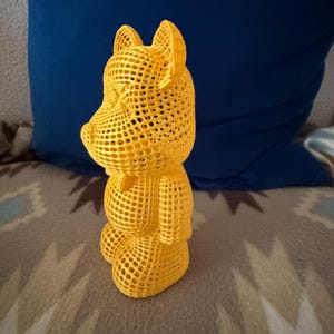 May include: A yellow, 3D-printed sculpture of a bear-like figure. The sculpture has a lattice-like design, with a hollow interior visible through the open mesh. The bear is standing upright, with its arms at its sides and its ears perked up.