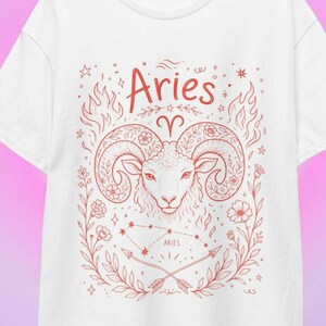 Könnte beinhalten: Ein weißes T-Shirt mit einer roten Linienzeichnung eines Widderkopfes in der Mitte, darüber steht das Wort Aries. Das Design umfasst florale Elemente, Flammen, Sterne, das Sternbild Widder und zwei gekreuzte Pfeile am unteren Rand des Drucks.