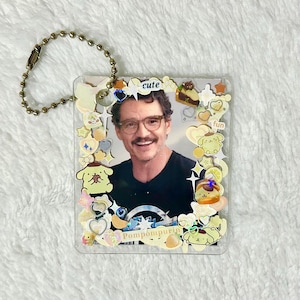 Peut inclure: Un porte-clés carré transparent avec une personne souriante portant des lunettes et un t-shirt noir. Le porte-clés est décoré de personnages de dessins animés, d'étoiles et du mot "cute". Une chaîne à billes dorée est attachée.