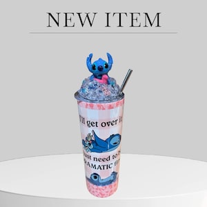 May include: A tumbler with a blue cartoon character figure on top, a silver straw, and the text "I'll get over it, just need to be dramatic first." The tumbler has a pink and white checkered pattern and a blue character graphic.