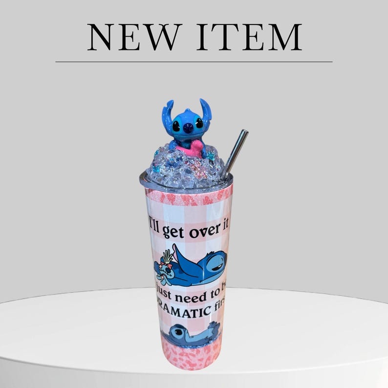 May include: A tumbler with a blue cartoon character figure on top, a silver straw, and the text "I'll get over it, just need to be dramatic first." The tumbler has a pink and white checkered pattern and a blue character graphic.