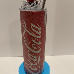 May include: A red glitter tumbler with a white Coca-Cola logo and a red cherry on top. The tumbler is sitting on a blue coaster.