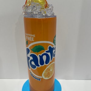 May include: Orange-colored stainless steel tumbler with a white lid and a printed image of a Fanta orange soda can. The can has the words "Fanta Orange" and "Caffeine Free" printed on it. The tumbler is filled with ice and topped with an orange slice.